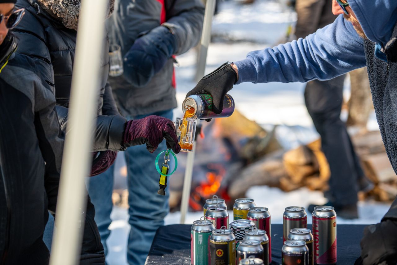 A beer vendor pours beer from a can into a patron's mini beer glass at an outdoor, winter event.