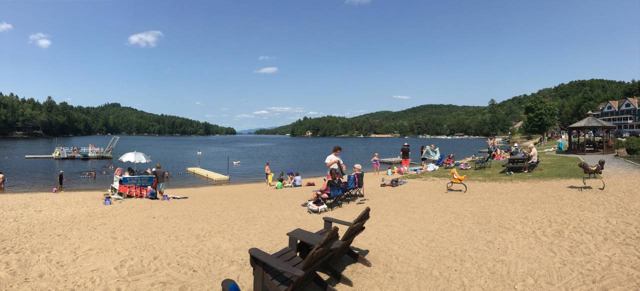 A small sandy beach on a sunny summer day. People lounge on the sand and a large, floating swimming dock is in the distance in the water.