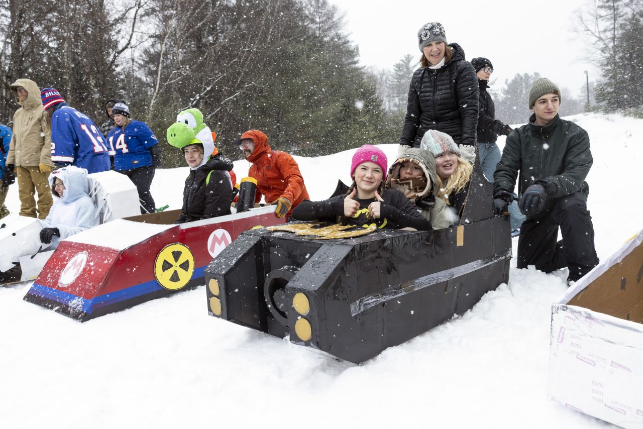 A group of children and adults pose in and next to two elaborately decorated cardboard sleds, one resembling a red Mario Kart and the other a black tank-like vehicle, at the Cardboard Sled Races at the Long Lake Winter Carnival.