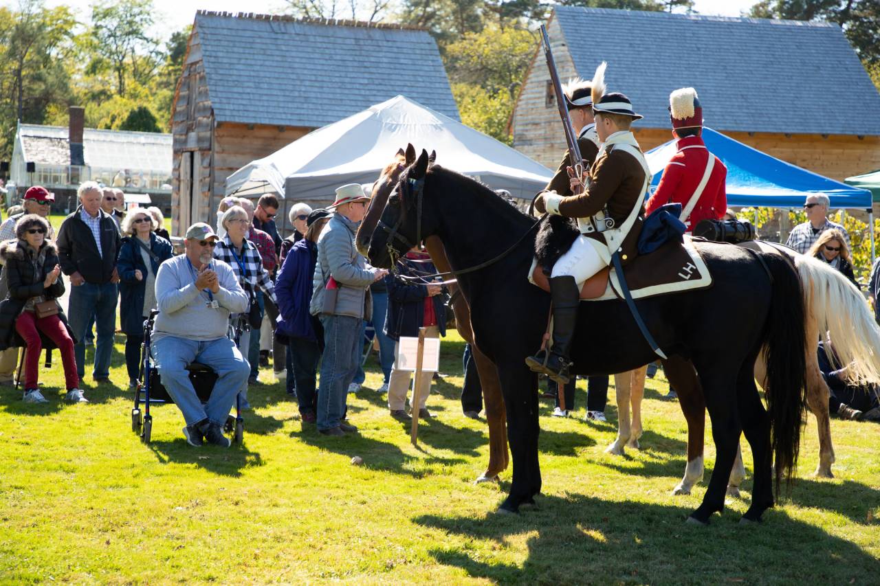 A group of older people watch as actors reenact Revolutionary war times on horseback. 