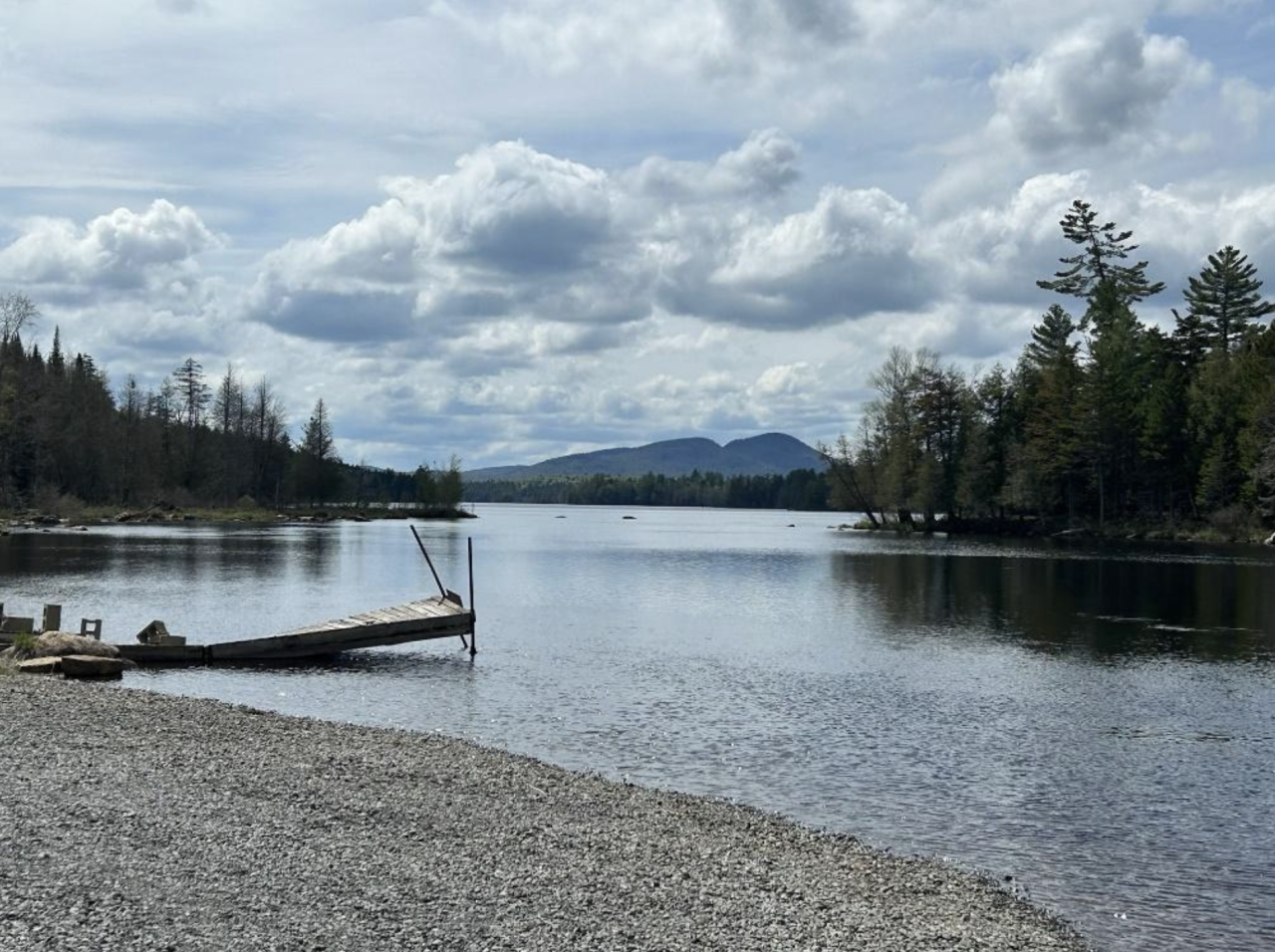 A view of a lake from a sandy beach. The lake is framed by evergreen trees.
