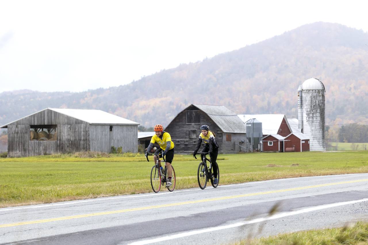 Two cyclists ride past a barn in fall. 