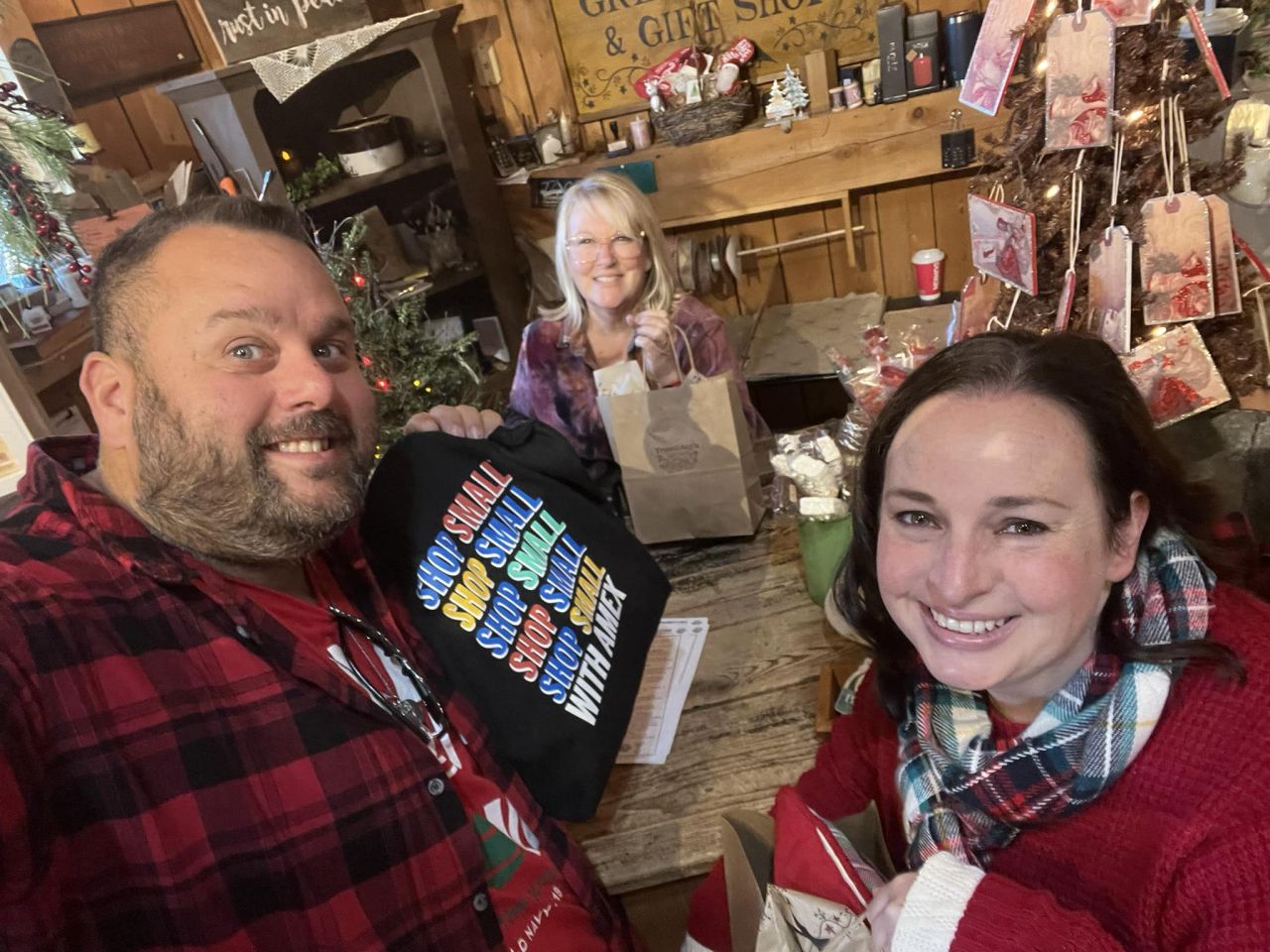 A man and woman dressed in winter clothes take a selfie with a store clerk in a gift shop. They're holding a bag that reads, "Shop Small"