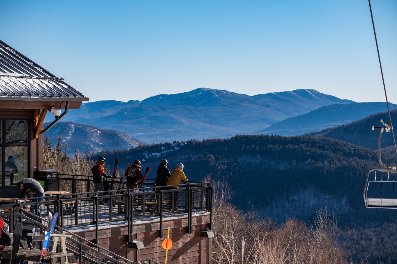 A group of skiers stand on an outdoor patio overlooking mountains. 