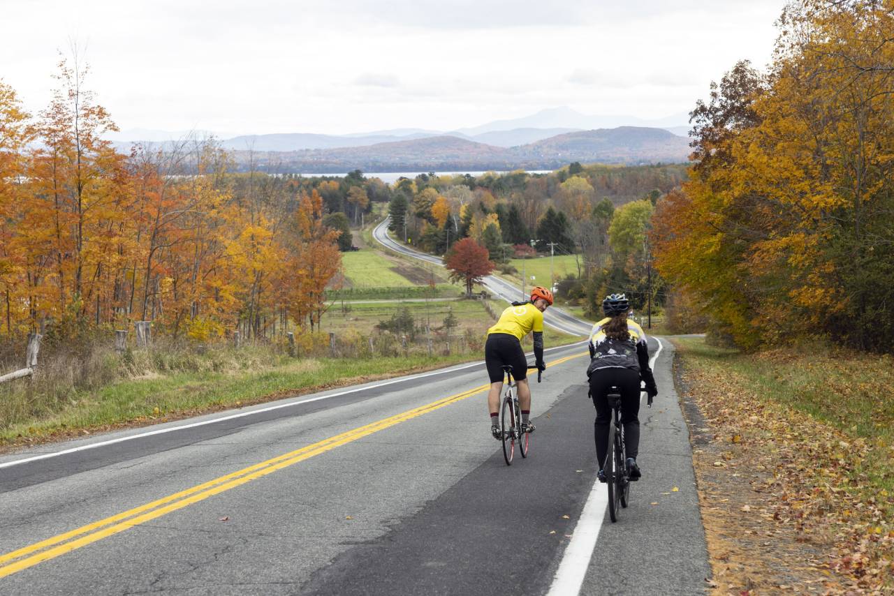Two cyclists ride down a hill in the countryside in fall. 