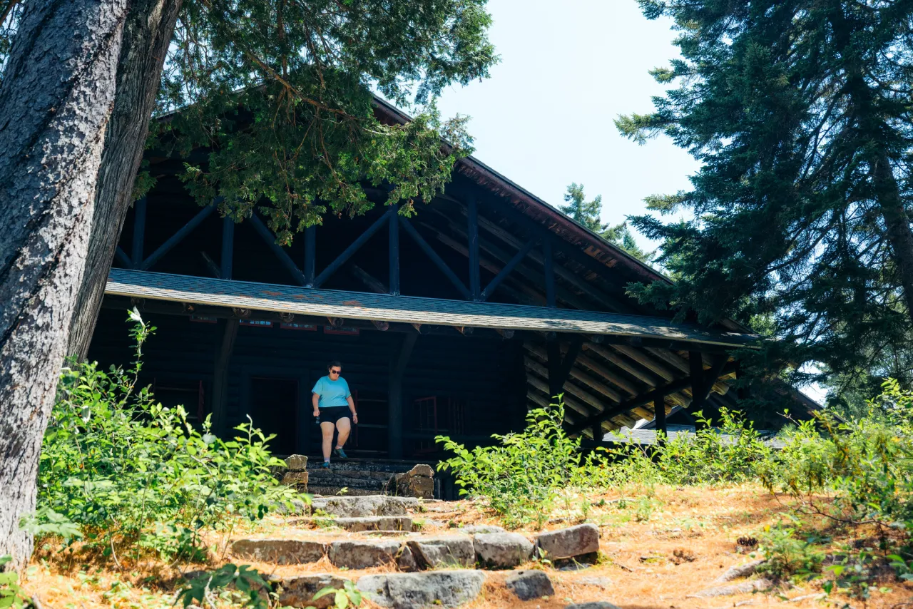 a woman walks down the steps of a log camp in the woods.