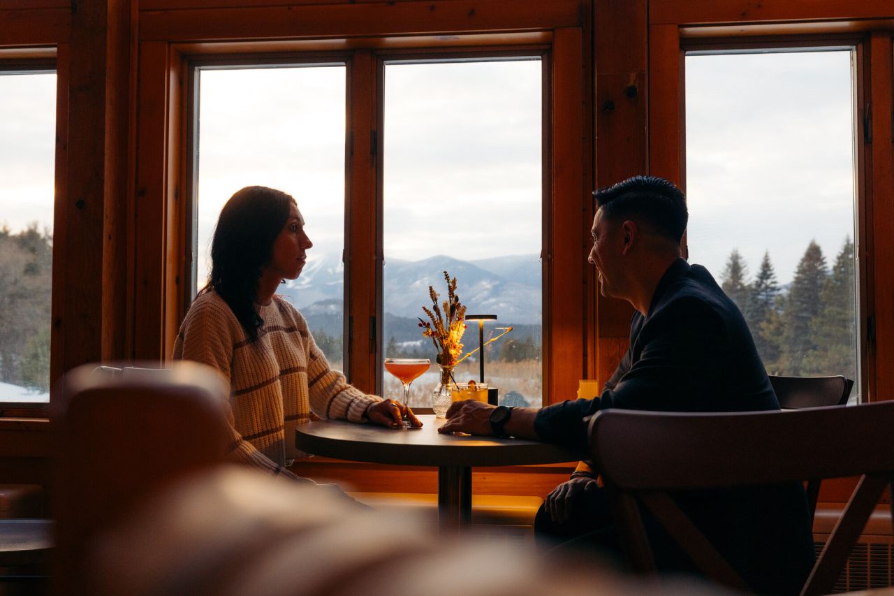 Man and woman sit across from another at a lamplit table with dried flowers and cocktails between them in front of large windows with winter mountain views