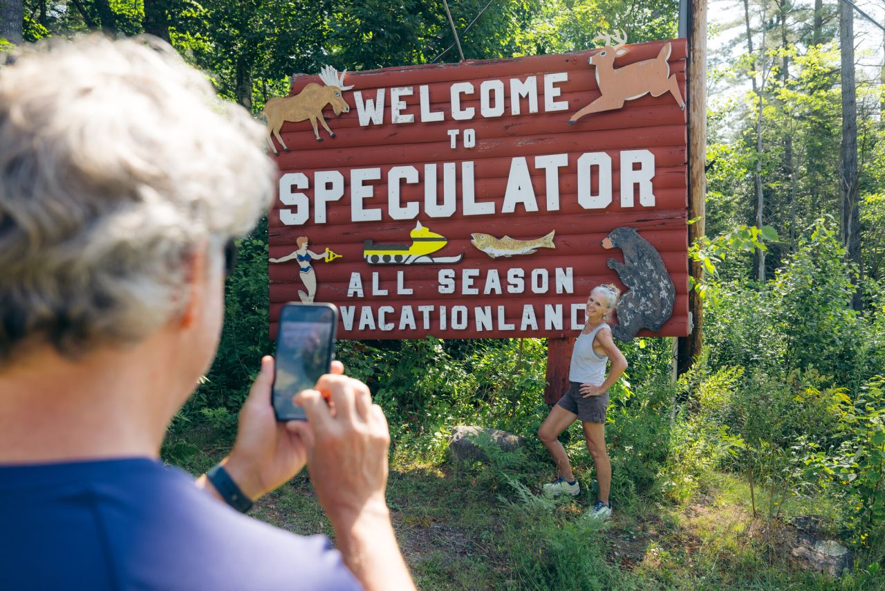 A man takes a picture of his wife in front of a town sign that says "Welcome to Speculator All Season Vacationland." 