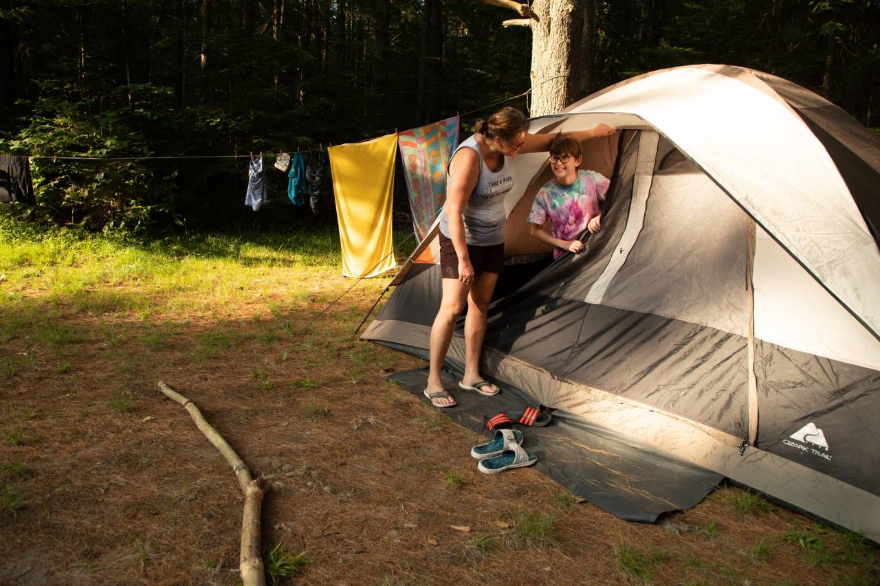 A woman and a child stand next to a tent on a pine needle-strewn campsite.