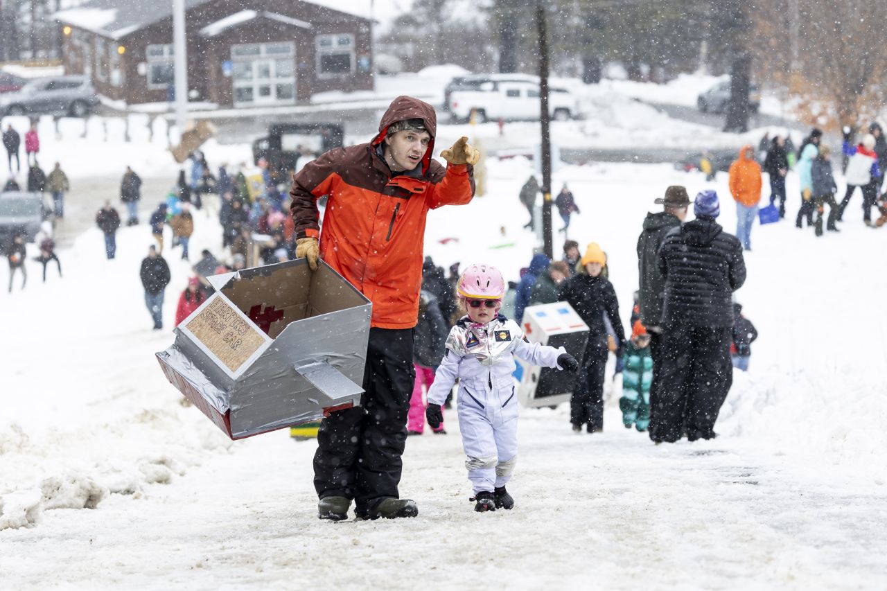 A man holds a gray cardboard box sled decorated to look like a house, walking next to a small child dressed in a white astronaut suit with pink helmet.