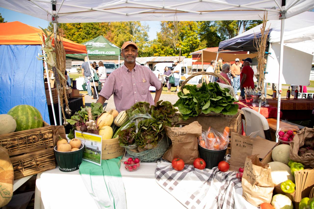 A man stands with a full table of harvested vegetables at a farmers market. 