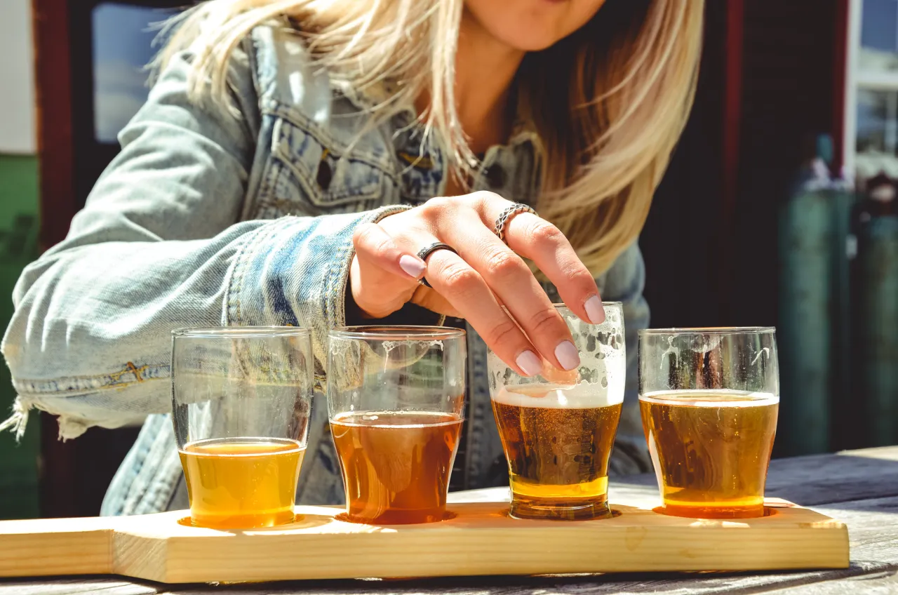 A close-up of a woman lifting a small glass of beer from a flight of beers on a sunny day.