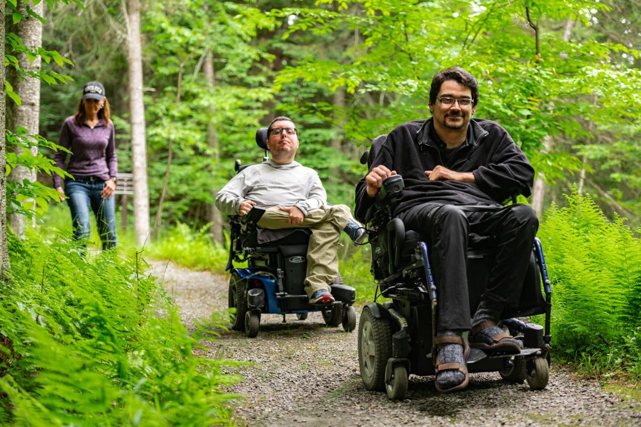 Two men in motorized wheelchairs  ride a gravel path. 