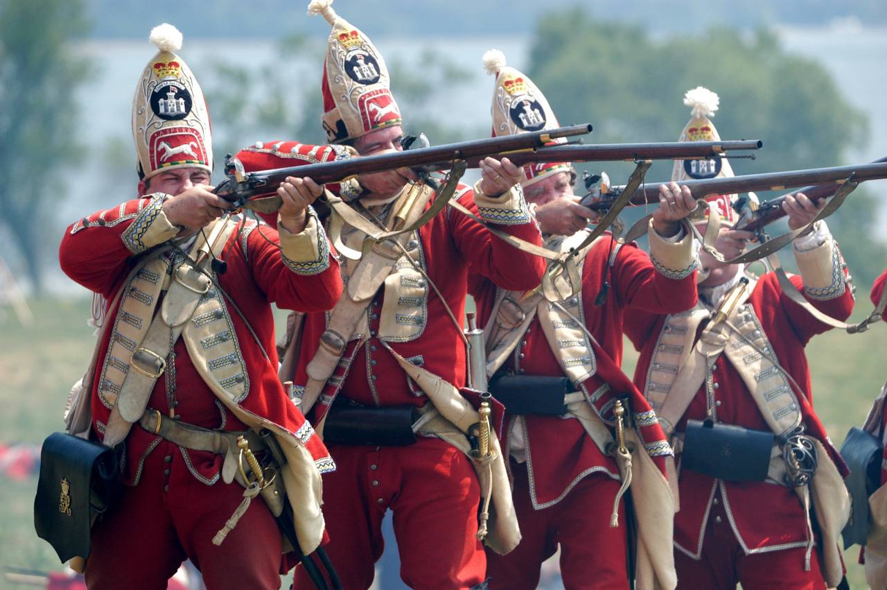 A team of red coat soldiers fire muskets. 