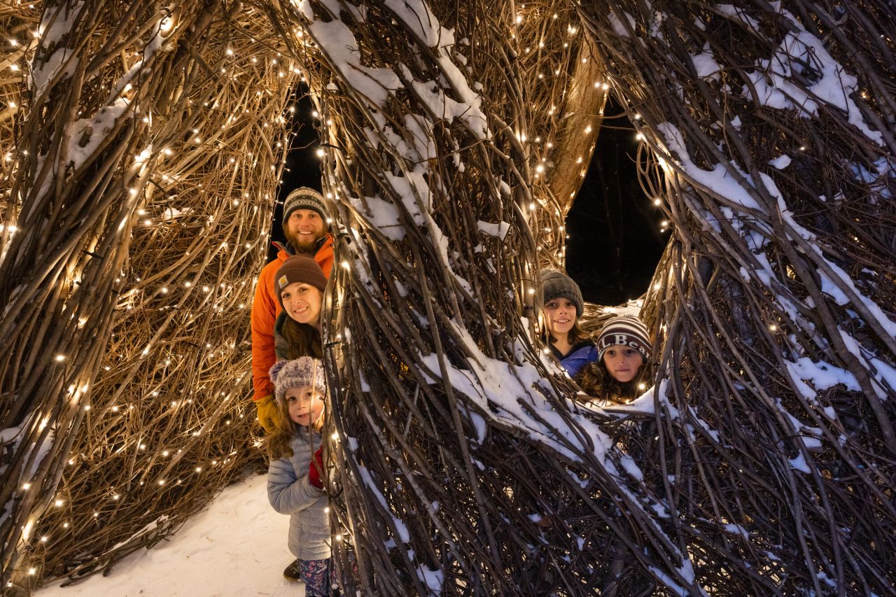 A family in winter gear poses for a picture around an art instillation of vines bound in lights.