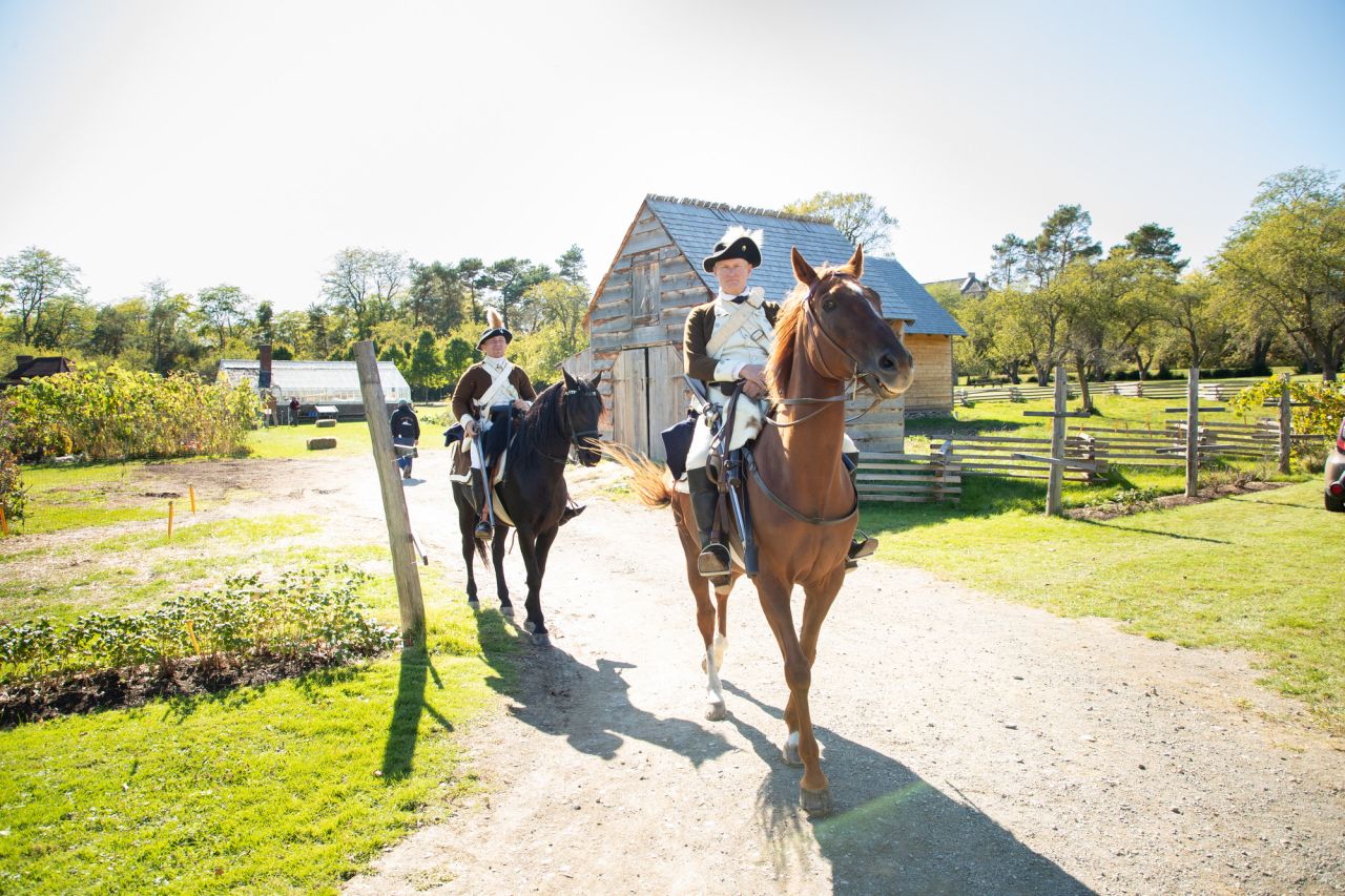 Two men in Revolutionary War costumes ride horses amid a rustic farm garden.