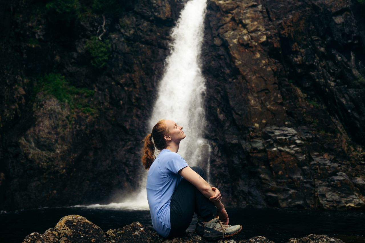 A woman sits and looks up at the sky in front of a waterfall. 