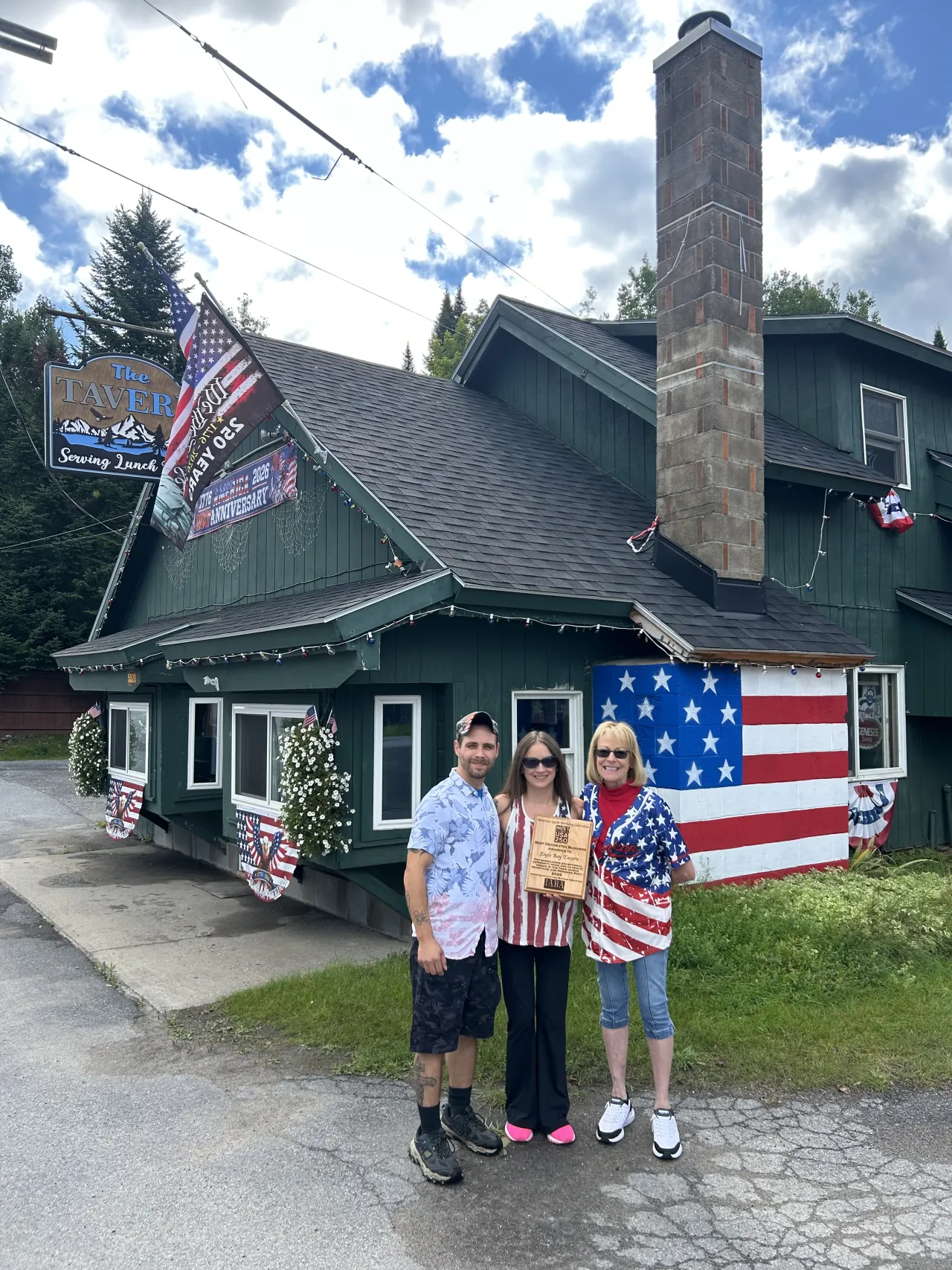 Three adults pose in front of a tavern decorated with American flags. They are holding an award.