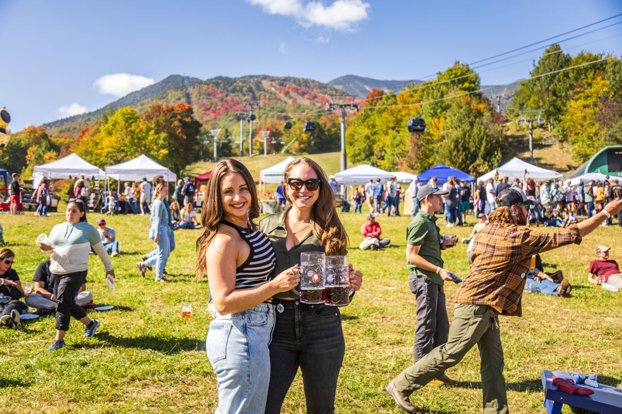 Two women smile holding mugs of beer at a fall event outside. 