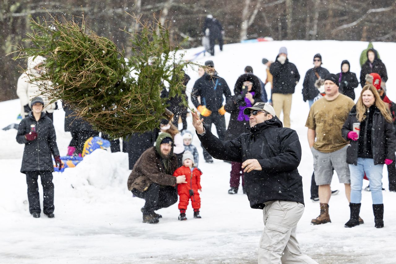 A group of spectators watches as a man wearing a baseball cap throws a large Christmas tree at the Christmas Tree Toss contest during the Long Lake Winter Carnival.