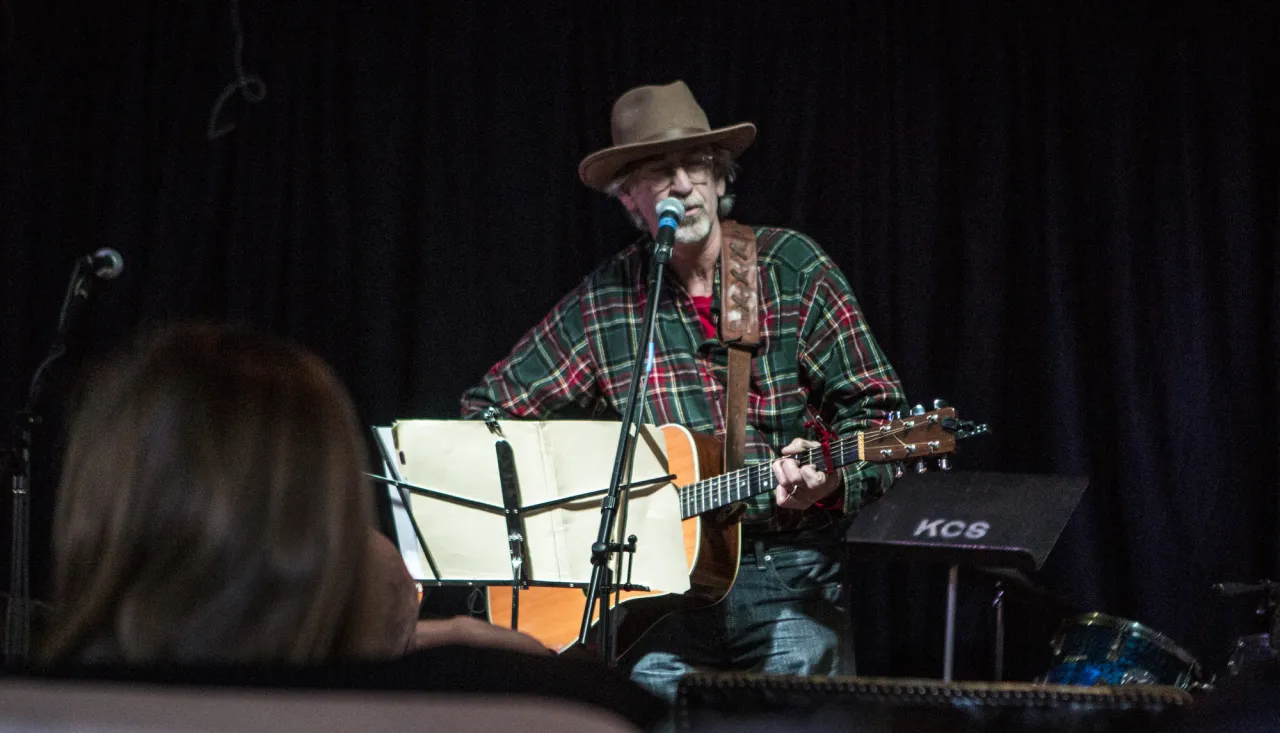 Man wearing wide brim hat plays guitar while singing into microphone seated behind music stand