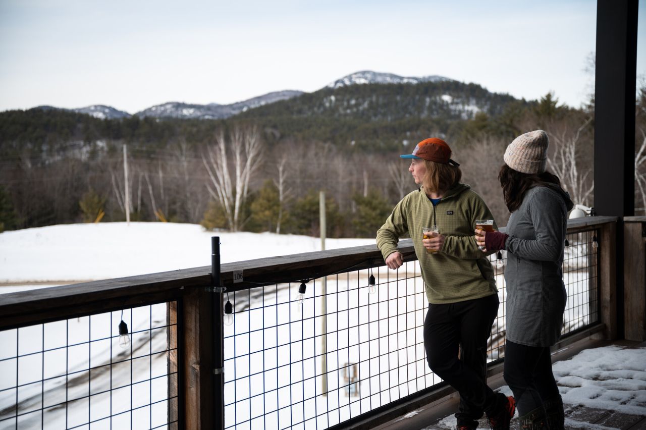 Two adults holding pint glasses of beer look out over a snowy field and hills.