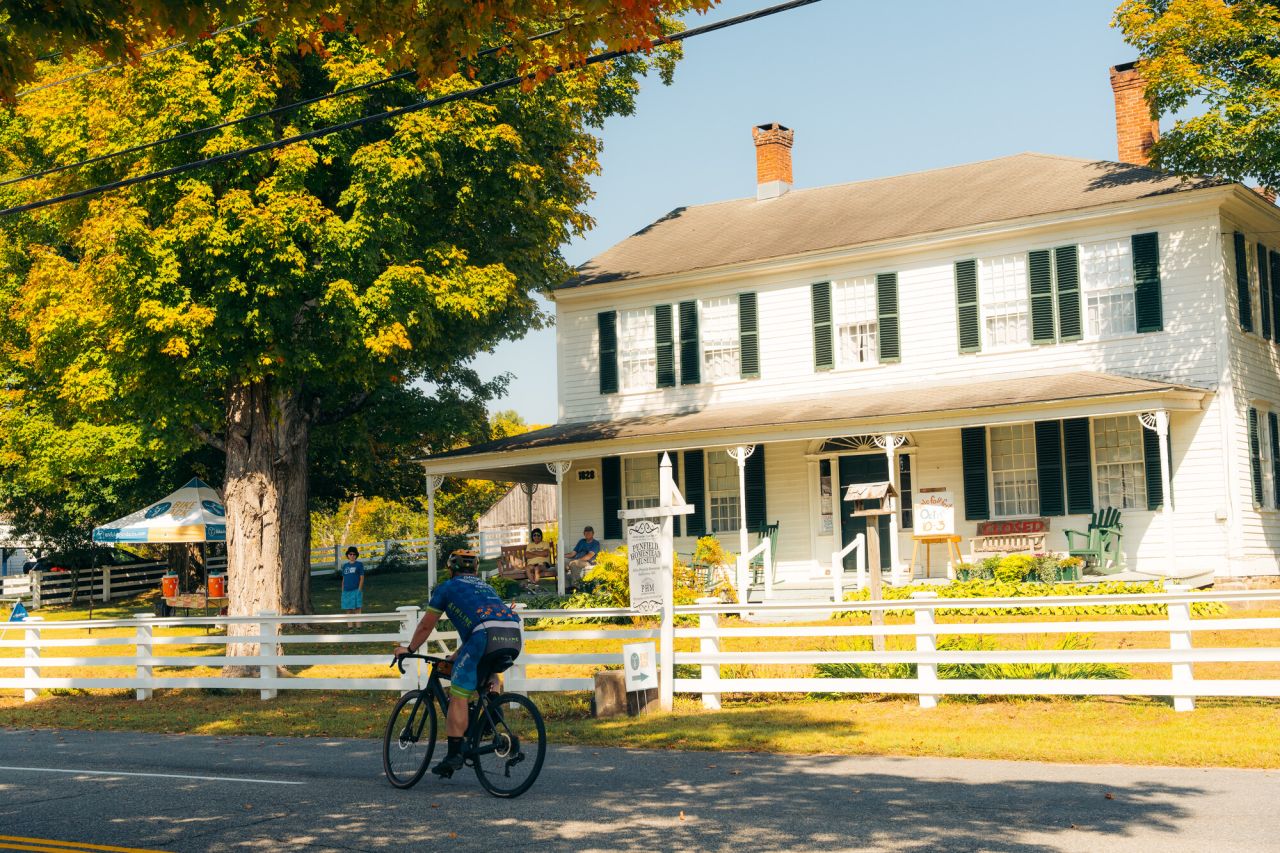 The exterior and front yard of a historic house museum. The building is a vintage Colonial home, white with green shutters and a broad front porch.