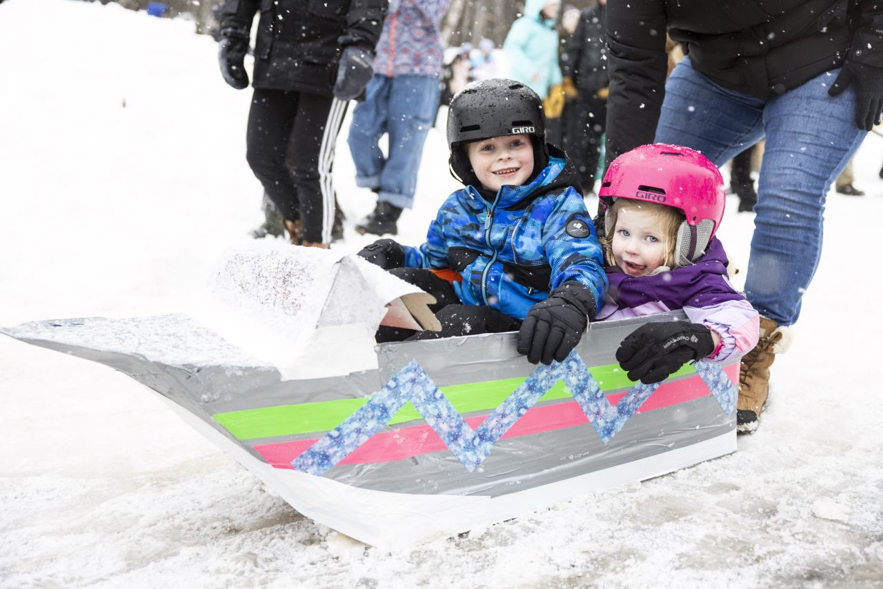 Two smiling young children in helmets and winter gear sit in a homemade cardboard sled designed like a boat.