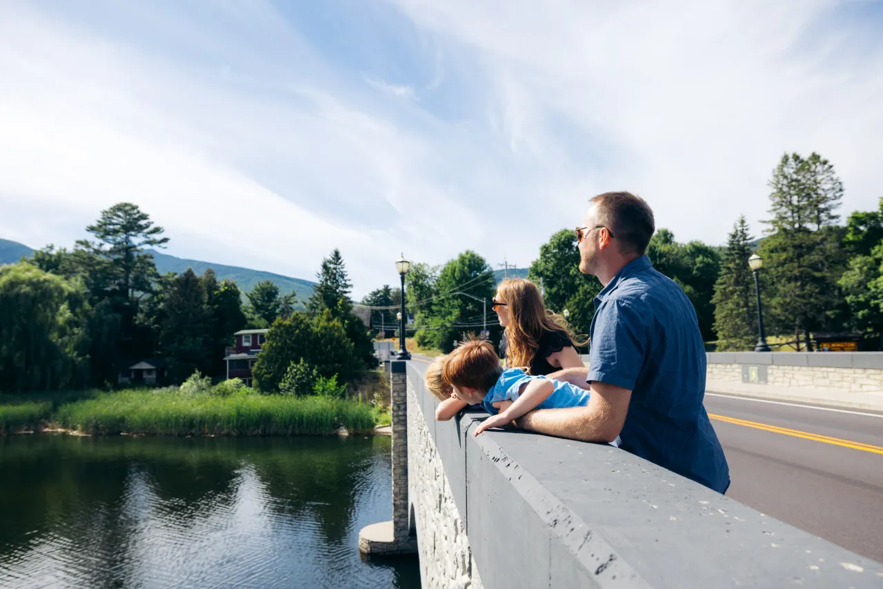 Parents stand on either side of young son and daughter on bridge over river looking out in summer