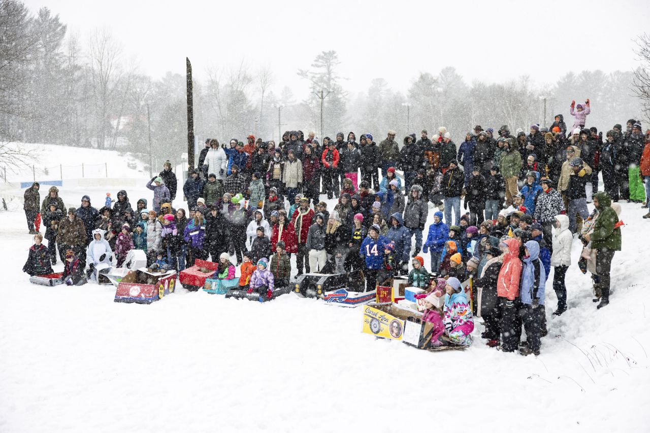 A large crowd of people in winter clothing stands on a snow-covered hill, posing for a group photo next to several decorated cardboard sleds, showcasing the turnout for the Cardboard Sled Races at the Long Lake Winter Carnival.