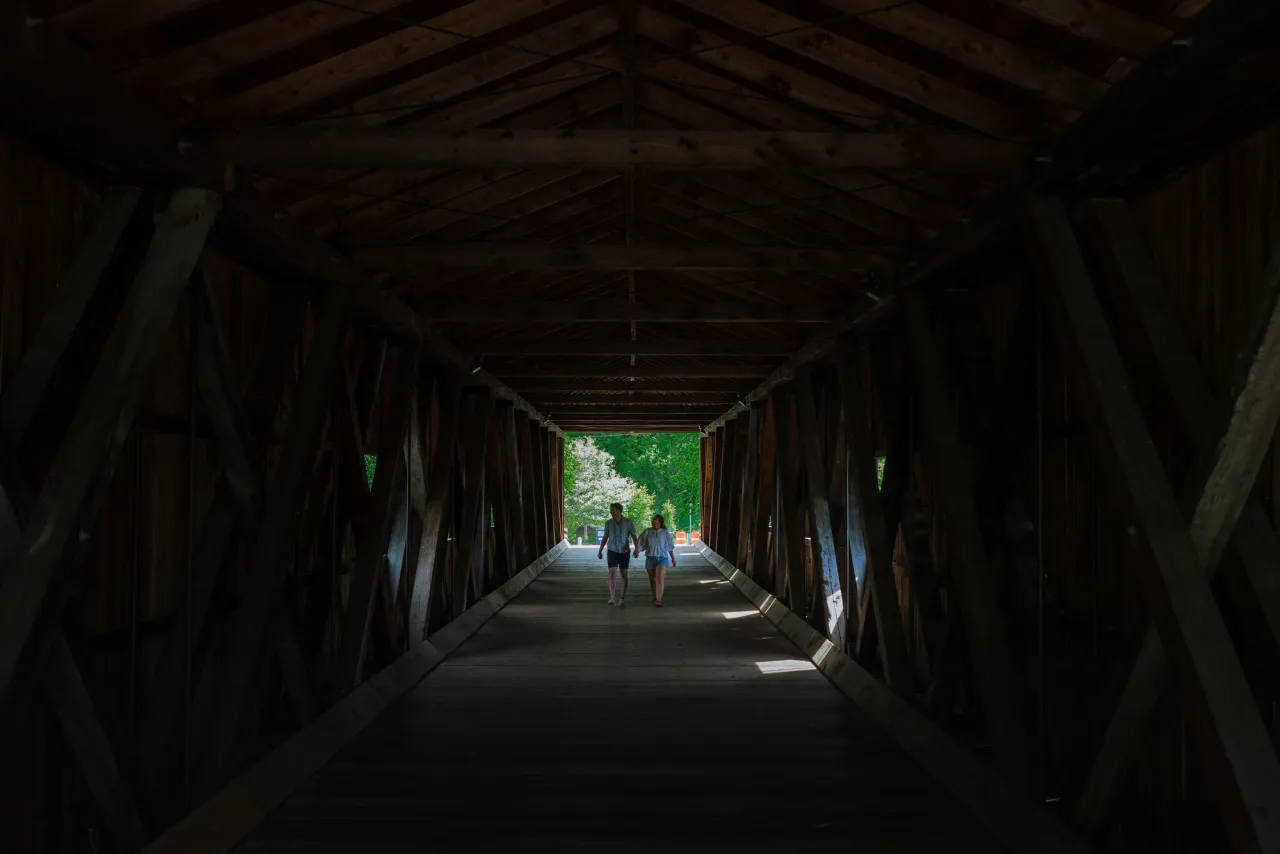 The dark interior of a wooden covered bridge with two people walking through the dark.