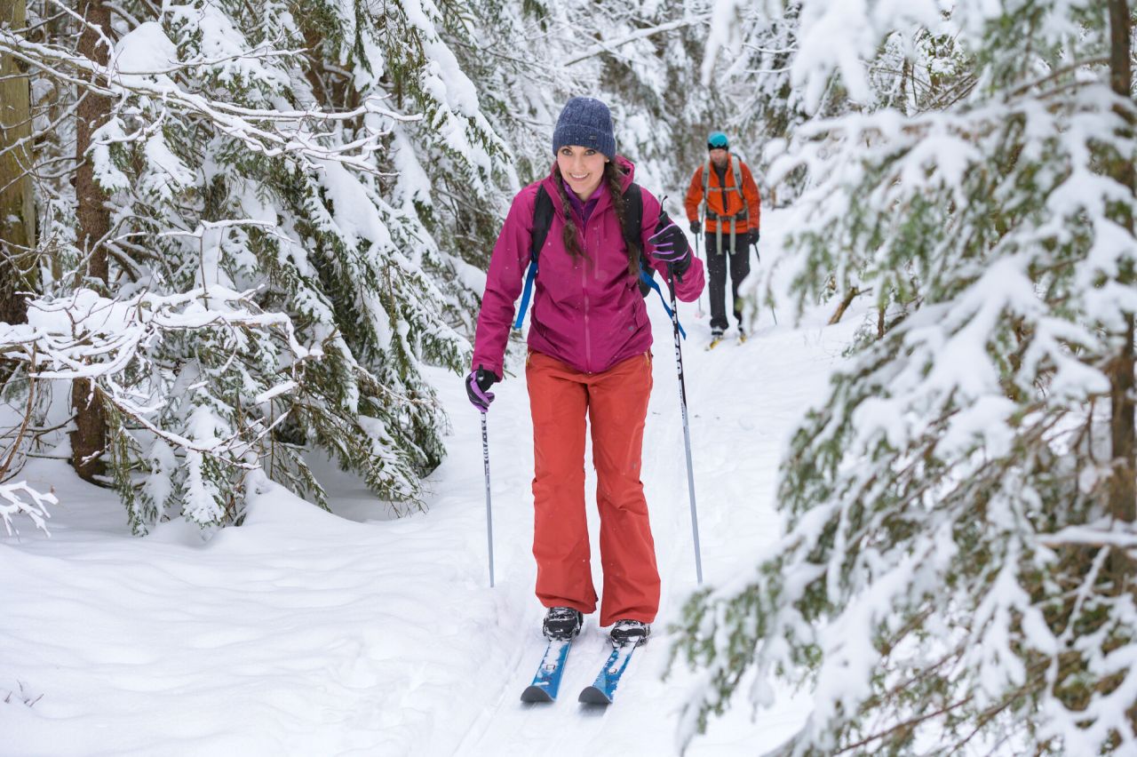 A woman cross-country skis in front of another skier on a snowy trail.
