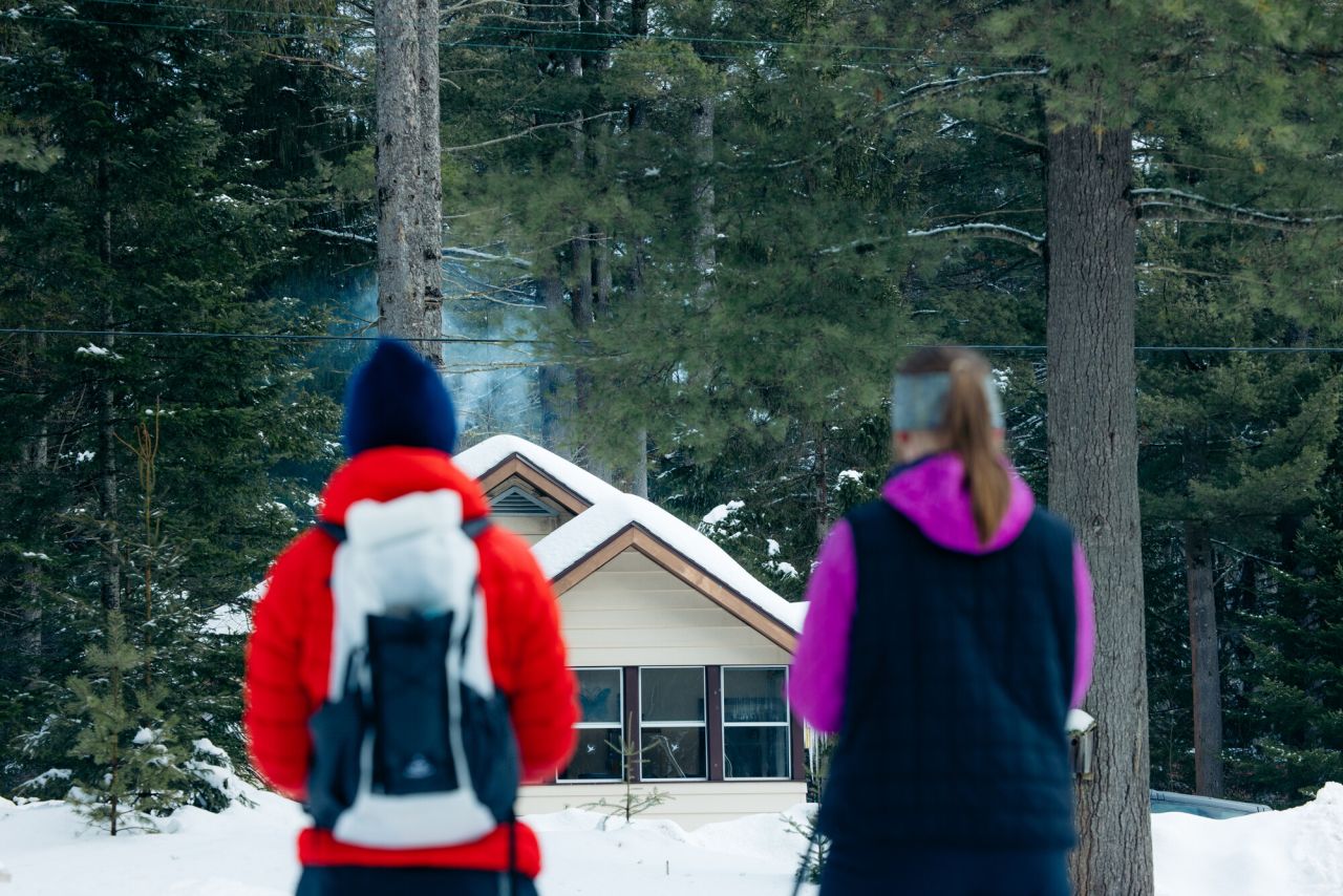 Two women on cross-country skis look out to a cottage