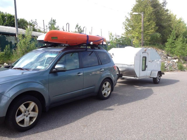 A blue SUV tows a white camper with an orange boat strapped to the roof. 