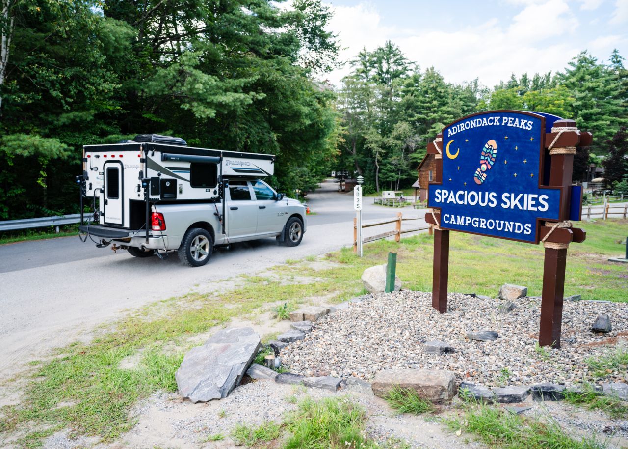 a truck hauling a camper drives past a sign for "spacious Skies Campground." 