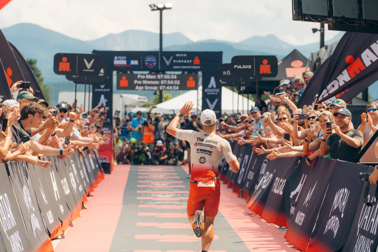 Ironman athlete waves as he runs chute flanked by people toward finish arch with High Peaks in the distant background