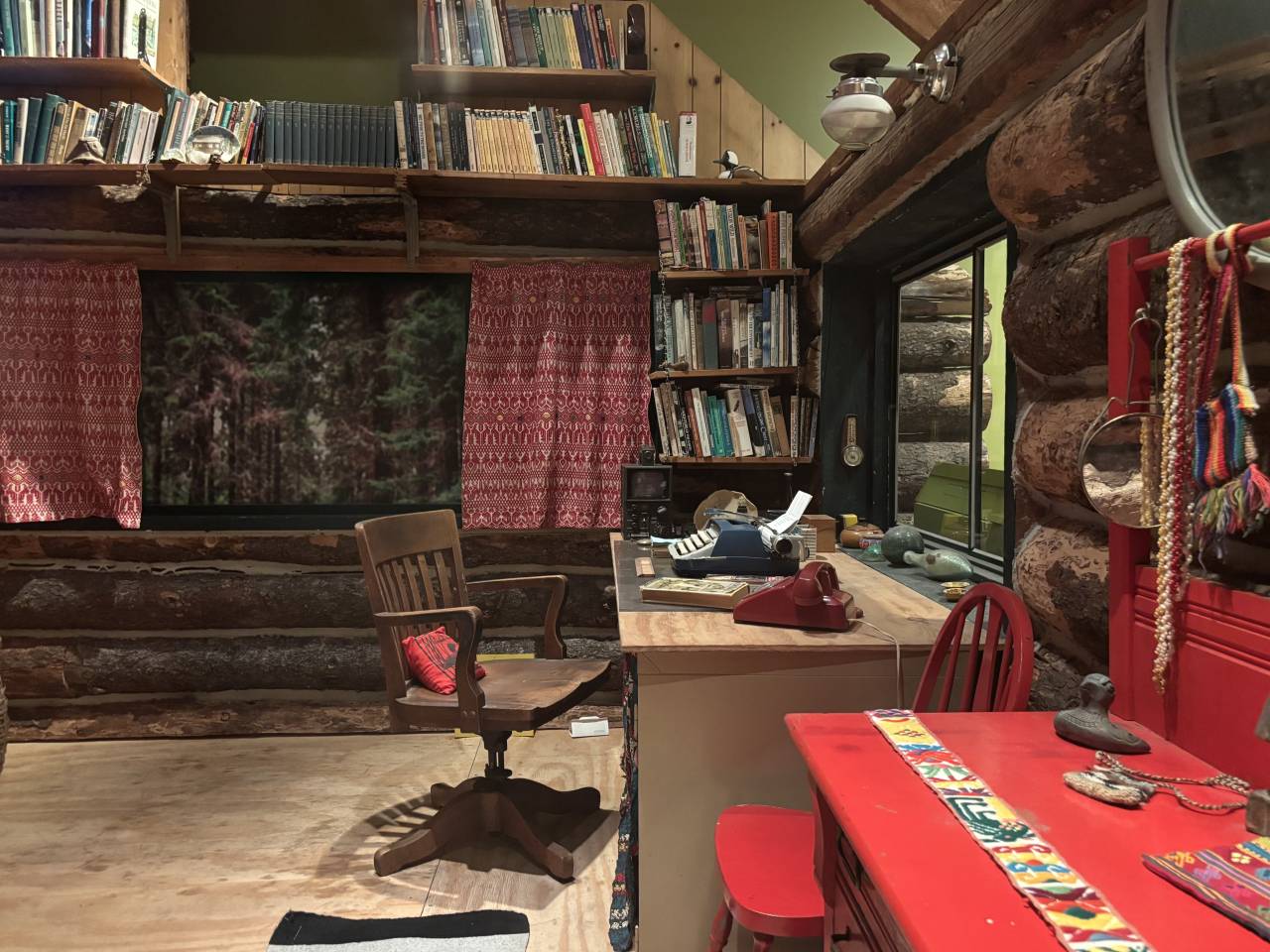 The interior of a rustic log cabin, including a desk with a typewriter on top, a small red dresser, and shelves of books.