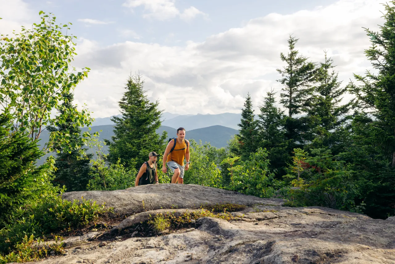 Two men are cresting a section of open rock face between mostly pine trees on a mountain top in summer