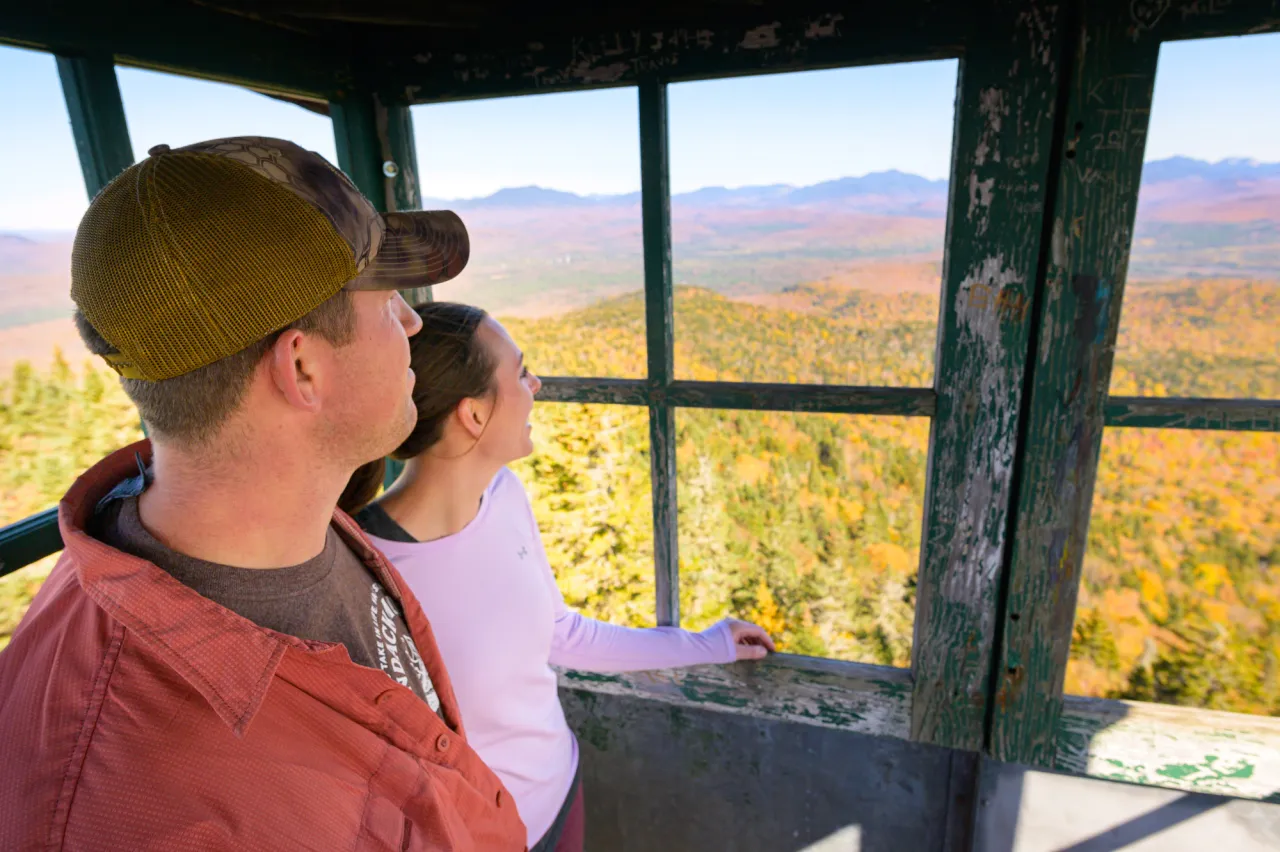 A man and a woman stand in a metal fire tower, looking out over a landscape of forest and mountains covered in fall foliage.