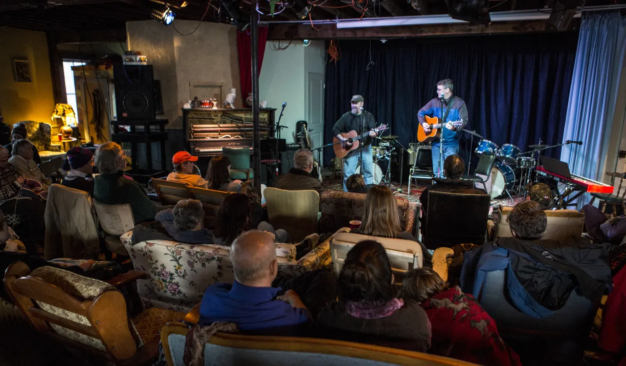 Two musicians play on Recovery Lounge stage while audience seated in mis-matched upholstered chairs look on