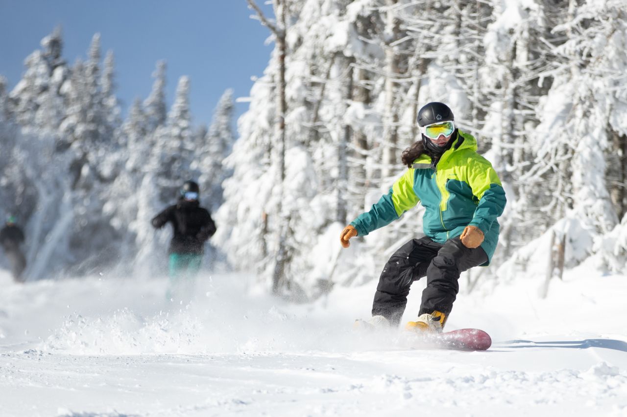 A snowboarder rides down a snowy slope on a sunny winter day with a snow-covered forest in the background.