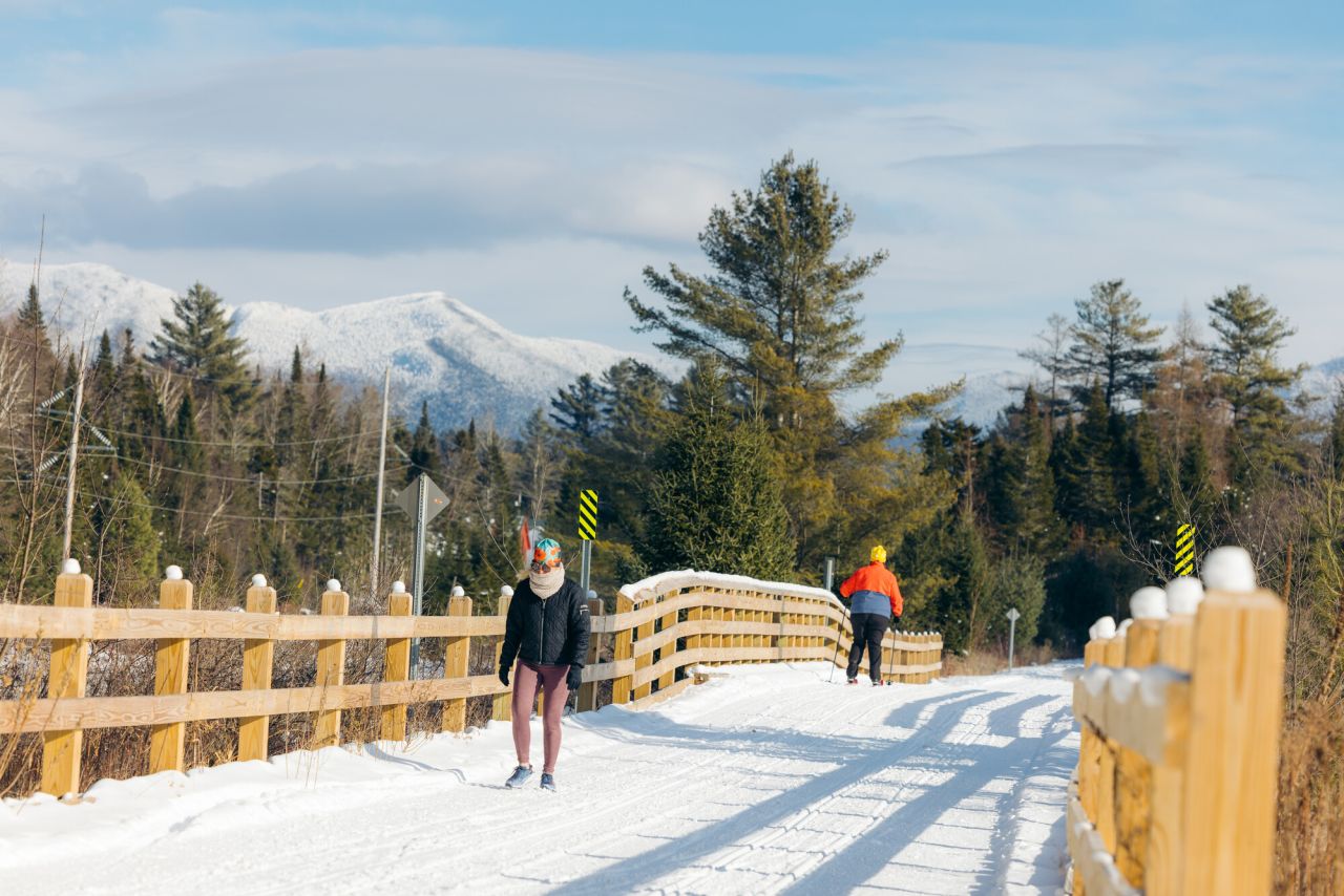 A woman walks past a cross-country skier on a  snowy fenced winter trail. 