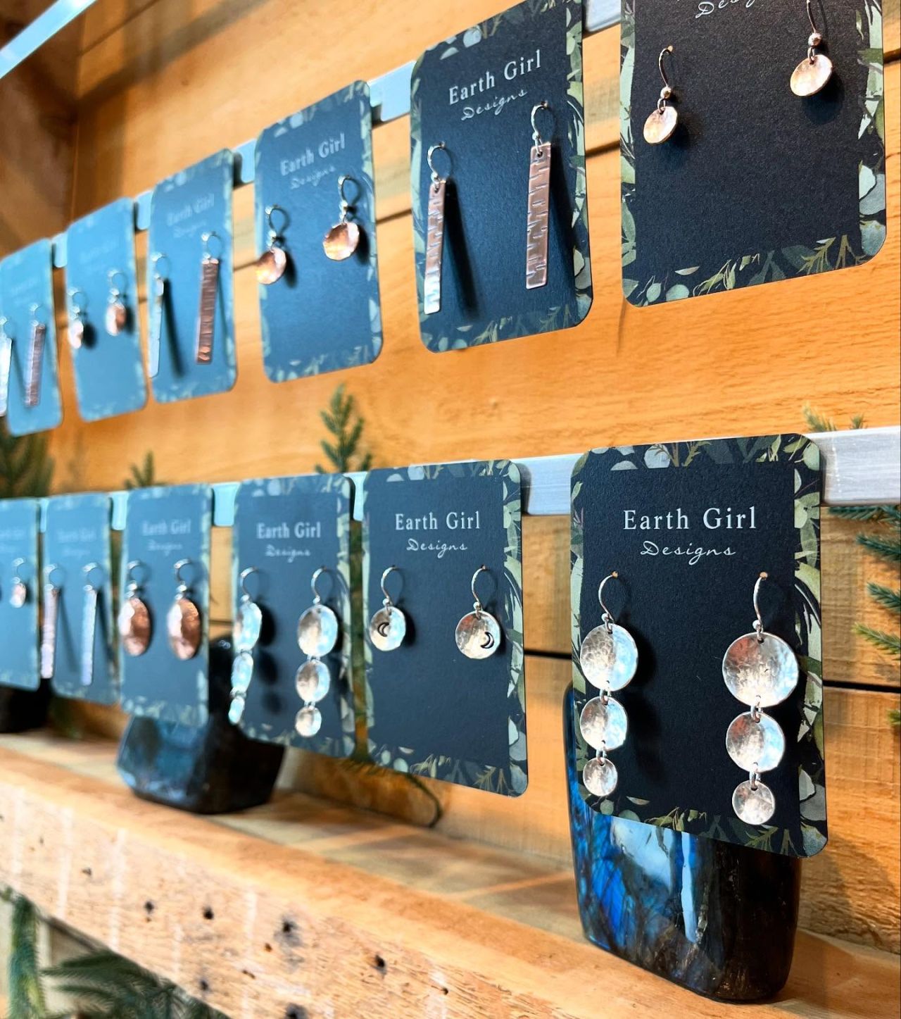 Earrings hang from small square display cards on two rows against a rustic wood backdrop.