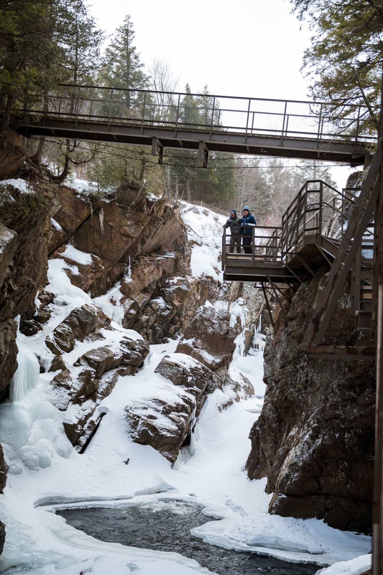 A man and woman look from a bridge down below to an ice covered waterfall. 