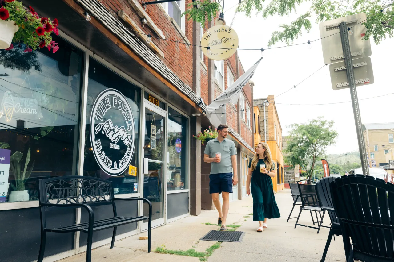 Man in shorts and polo and woman in dark dress walk on sidewalk holding cold coffee drinks past Forks Blend and Brew