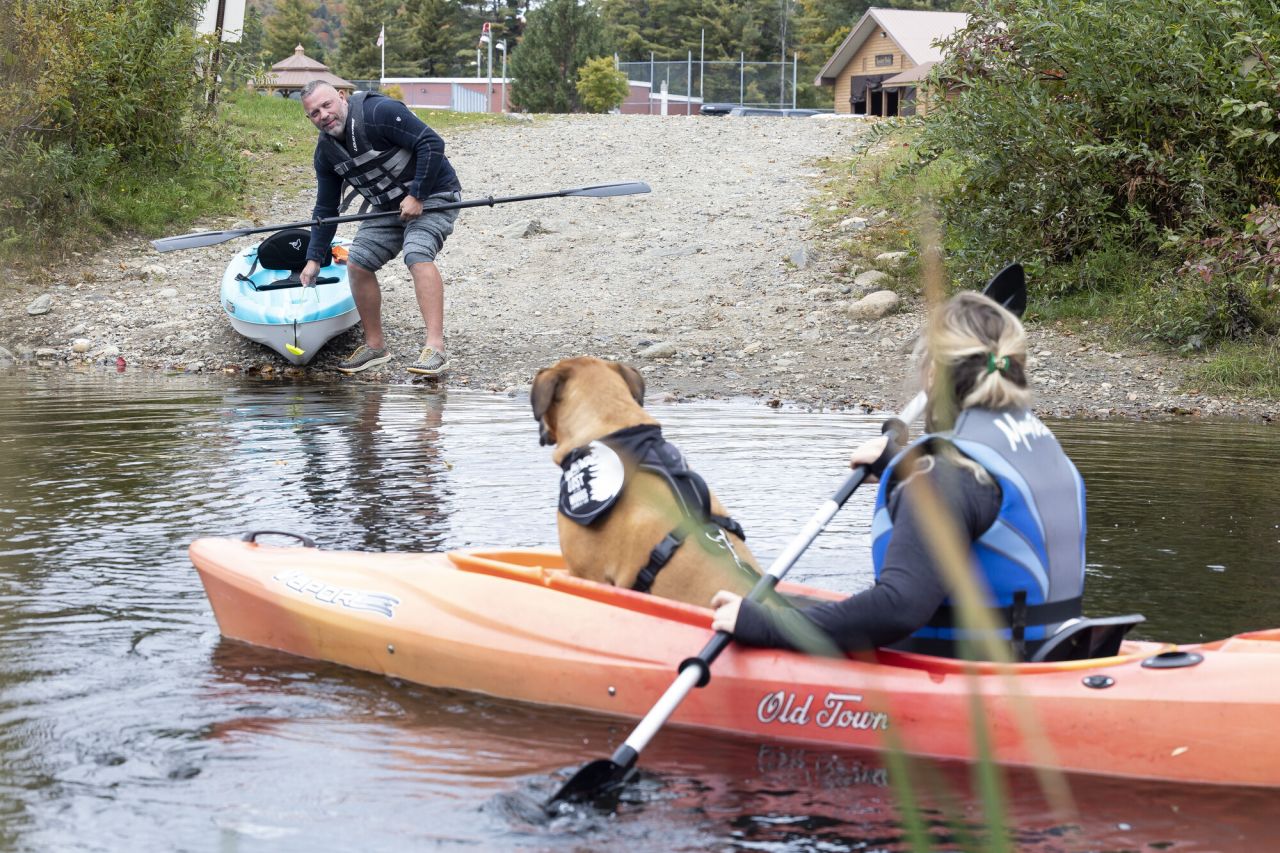 A man puts a kayak into the water of a lake while a woman sits in a kayak with a dog in front  of her. 
