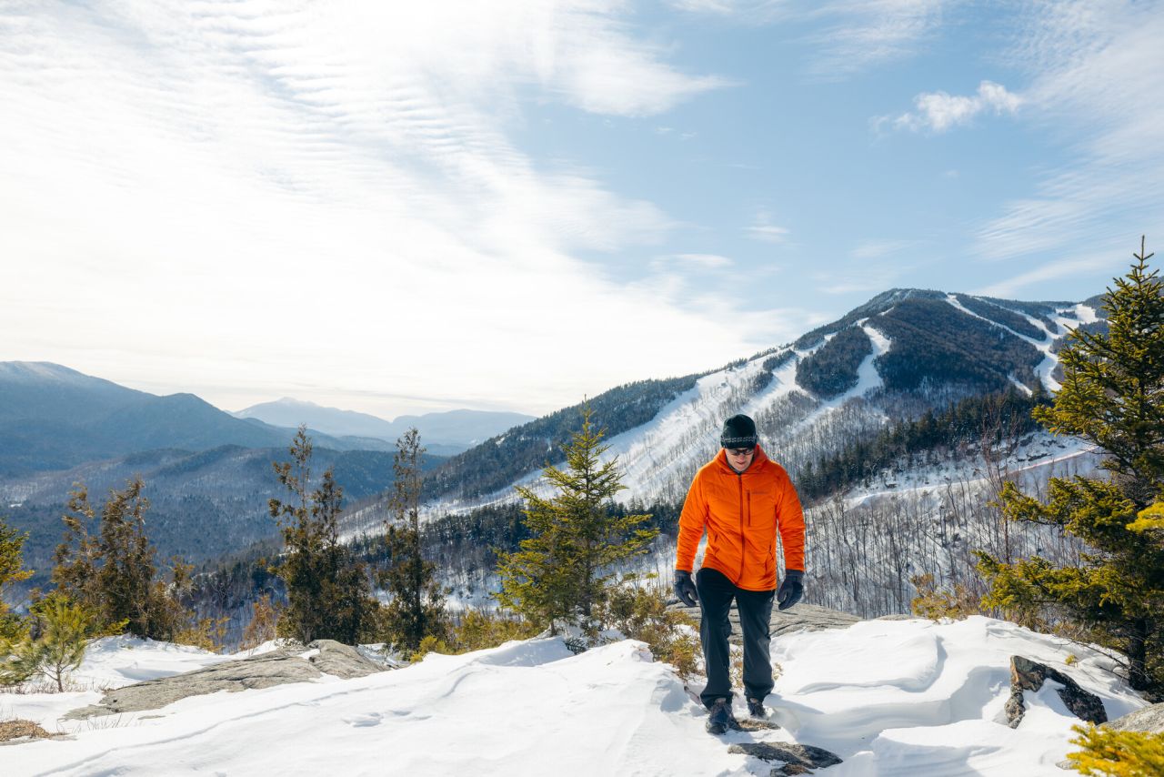 Hiker in bright orange coat and winter gear walks along snowy mountain summit with Whiteface Mountain in the background on sunny winter day