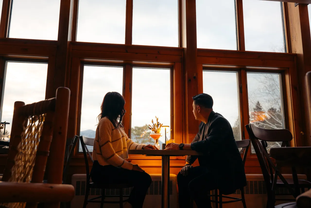 Woman and man sit opposite each other at table in front of floor-to-ceiling window with cocktails and flowers on table