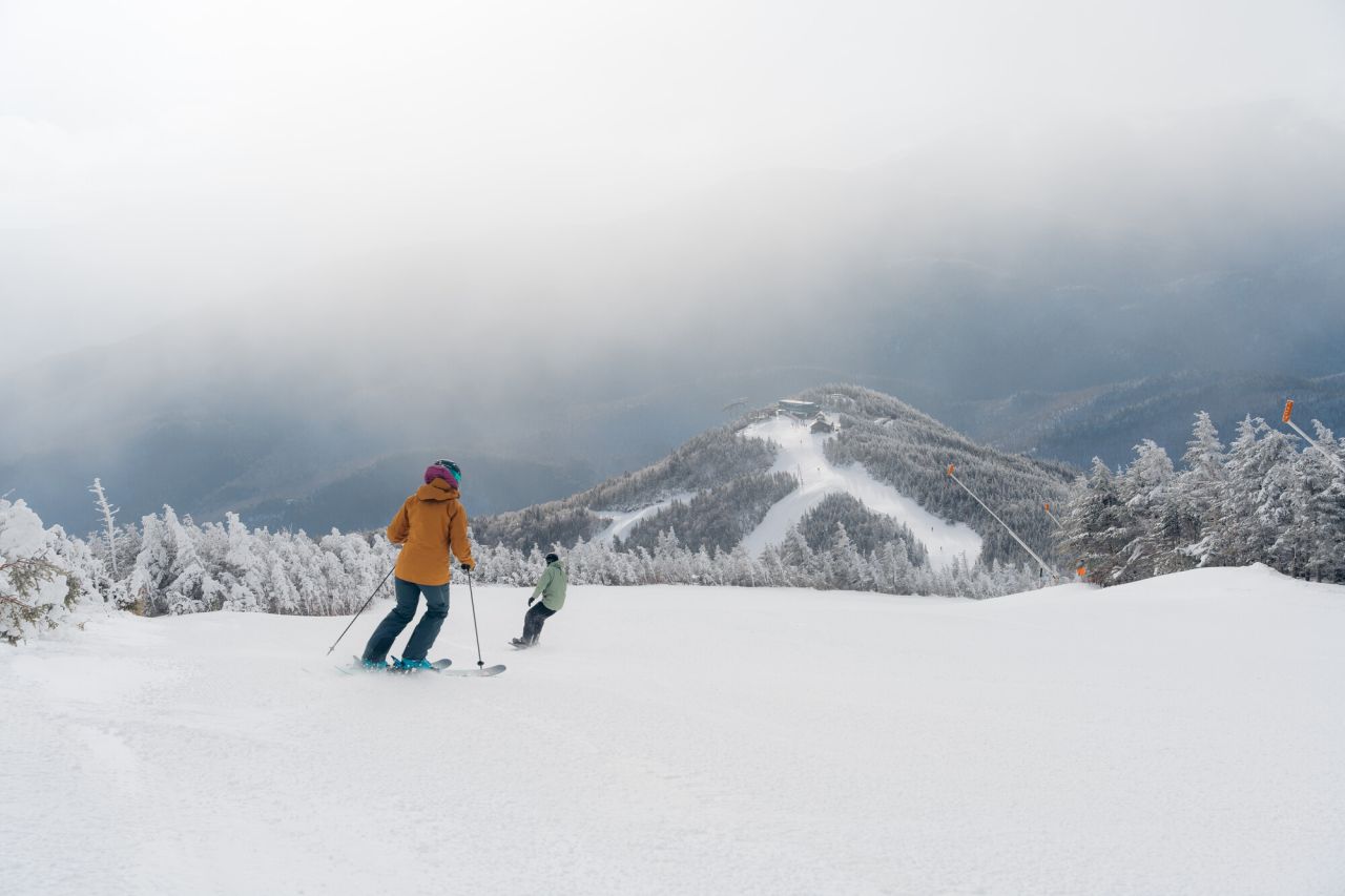 Skier and snowboarder descend one after the other on a wide open Whiteface Mountain ski trail with a smaller mountain summit with ski lines visible below