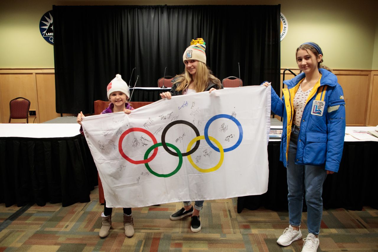 Three kids hold an Olympic flag while wearing vintage 1980 Olympic gear.